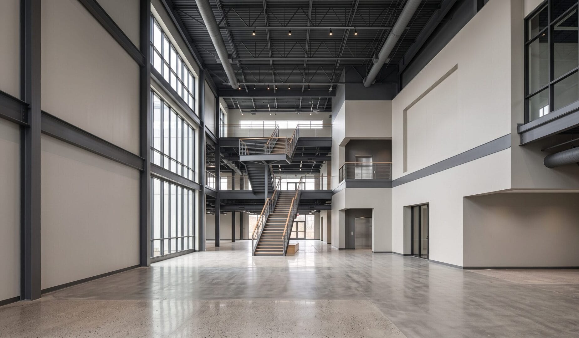 Spacious modern building interior with high ceilings, large windows, exposed beams, a central staircase, polished concrete floors, and multiple levels with open hallways and glass-walled offices.