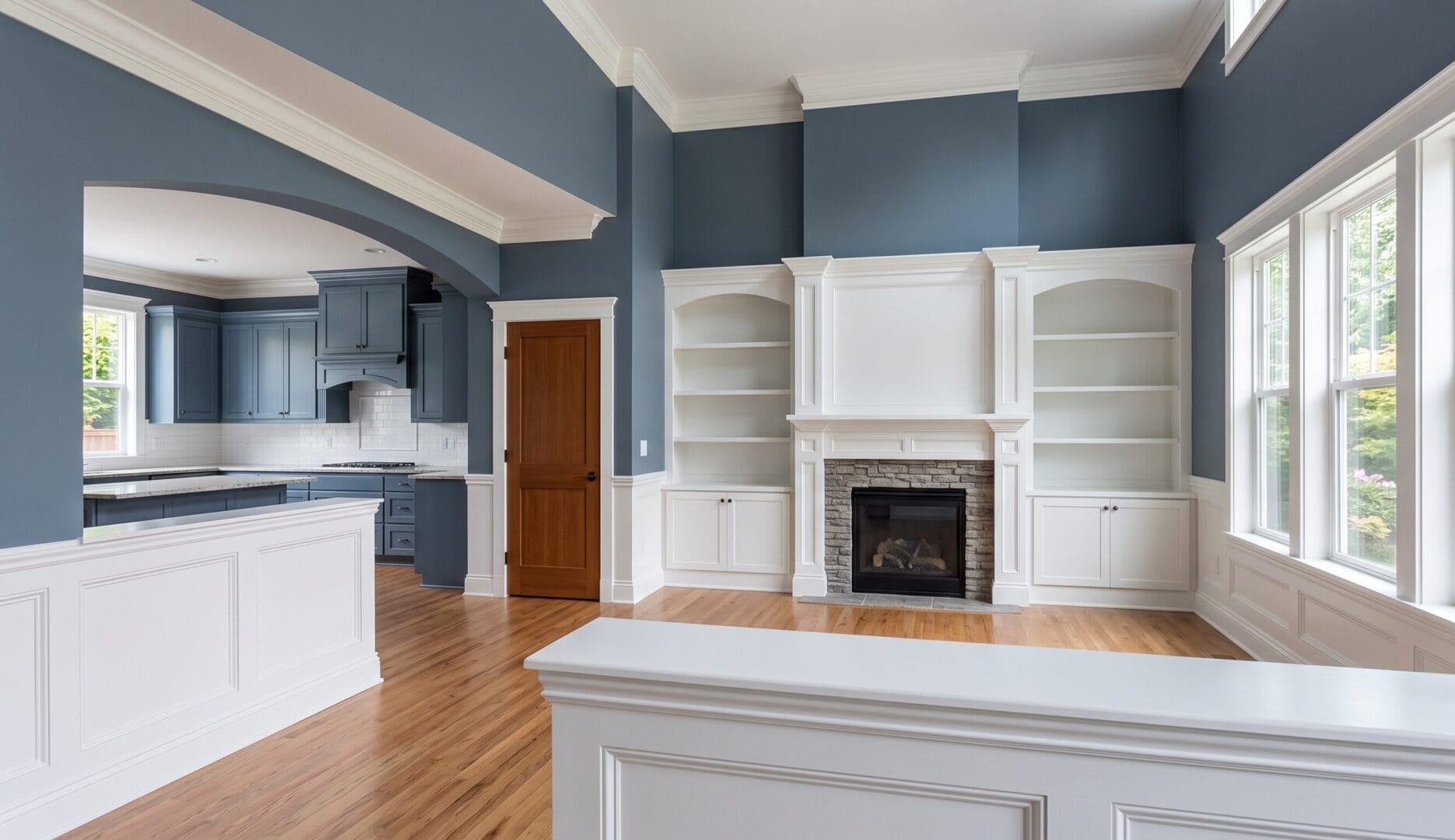 Spacious living room with blue walls, white trim, built-in shelves, a fireplace, hardwood floors, and large windows. Kitchen with blue cabinets visible through an arched opening.