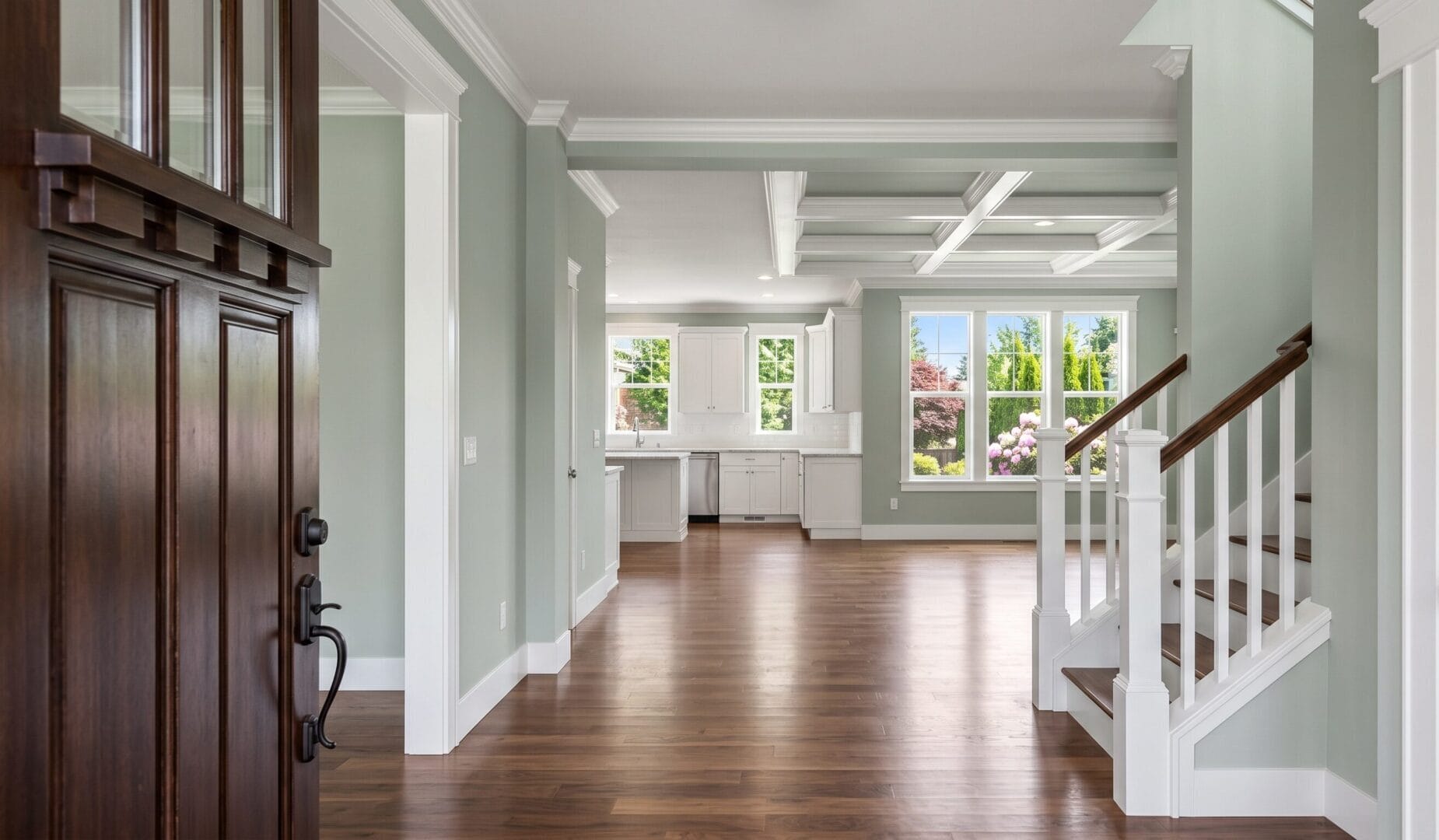 A bright, empty home interior with wood floors, light green walls, white trim, an open kitchen, large windows, and a staircase with white spindles and a wooden handrail.