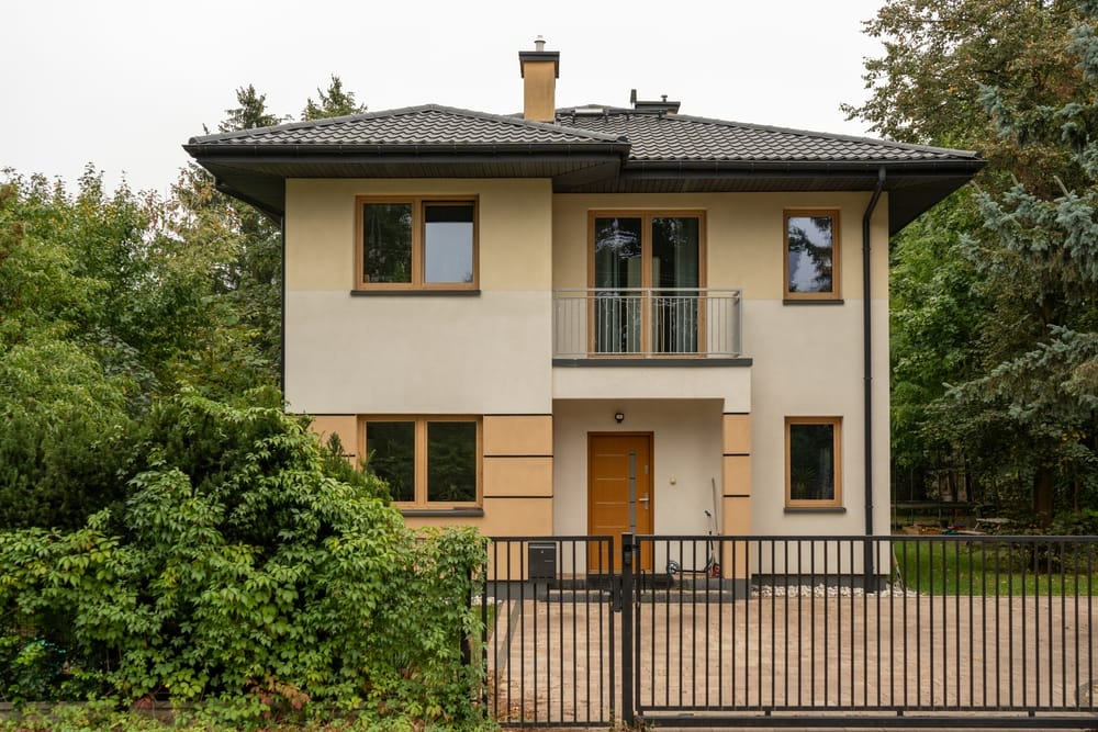Modern two-story house with cream and beige exterior, flat dark roof, a small balcony, and large windows. Surrounded by trees and greenery, with a black metal gate and brick driveway in front.
